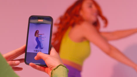 Studio-Shot-Of-Woman-Taking-Photo-Of-Friend-Dancing-On-Mobile-Phone-Against-Pink-Background-7